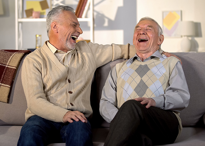 Bild: Zwei ältere Männer sitzen auf einem Sofa; beide lachen voller Inbrunst. Der linke Mann sitzt dem anderen Mann zugewandt, ein Arm auf der Rückenlehne. Die Stimmung wirkt vertraut und ausgelassen.