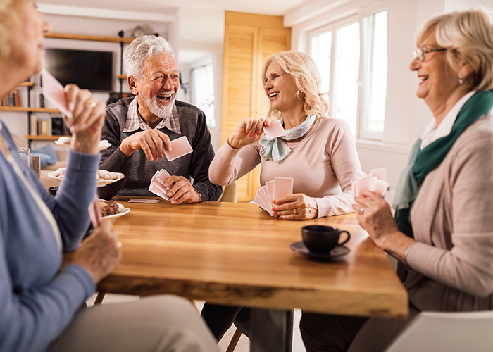 Bild: Vier ältere Menschen sitzen an einem Tisch. Sie spielen Karten und lachen sich an. Auf dem Tisch steht eine Kaffeetasse und eine Etagere mit Keksen. Die Stimmung wirkt verspielt und vertraut.
